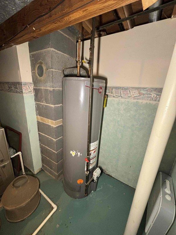 Provided estimate for complete water heater replacement in basement utility area. Assessed existing gray cylindrical water heater with copper and black piping connections, evaluated current plumbing configuration including overhead pipe routing through floor joists, and documented installation conditions in unfinished basement space with concrete block walls and turquoise-painted concrete floor for accurate replacement quote.