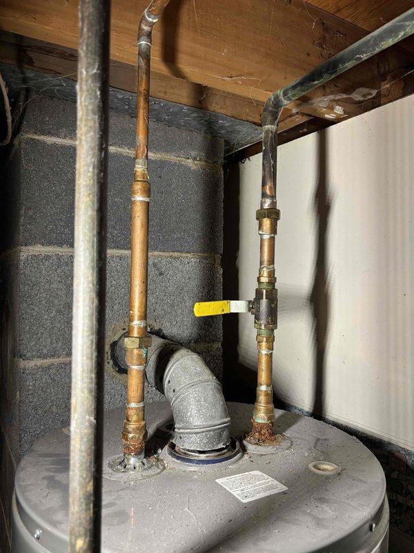 Provided estimate for complete water heater replacement in basement utility area. Assessed existing gray cylindrical water heater with copper and black piping connections, evaluated current plumbing configuration including overhead pipe routing through floor joists, and documented installation conditions in unfinished basement space with concrete block walls and turquoise-painted concrete floor for accurate replacement quote.