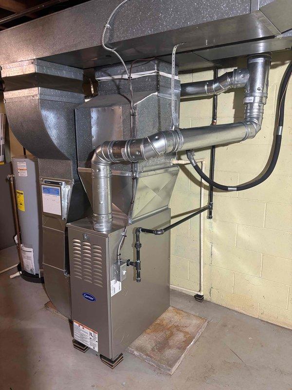 Completed full furnace installation in basement utility area, including Carrier-branded furnace unit positioned on concrete floor with comprehensive metallic ductwork system. Installed and secured extensive network of supply ducts extending from furnace to ceiling-mounted plenum, utilizing multiple elbow joints and straight sections to create complete ventilation pathway. Mounted air handler assembly near ceiling with proper structural support using black metal straps and hangers. Connected flexible ducting to upper portions of system and ensured all transitions were properly sealed. Verified all ductwork connections, tested system operation, and confirmed proper airflow throughout installation.