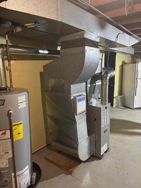 Completed full furnace installation in basement utility area, including Carrier-branded furnace unit positioned on concrete floor with comprehensive metallic ductwork system. Installed and secured extensive network of supply ducts extending from furnace to ceiling-mounted plenum, utilizing multiple elbow joints and straight sections to create complete ventilation pathway. Mounted air handler assembly near ceiling with proper structural support using black metal straps and hangers. Connected flexible ducting to upper portions of system and ensured all transitions were properly sealed. Verified all ductwork connections, tested system operation, and confirmed proper airflow throughout installation.