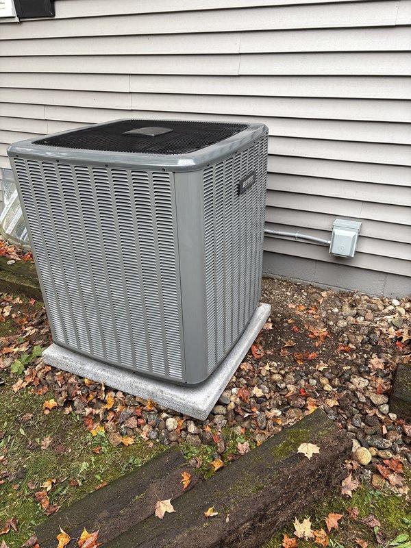 Completed full residential air conditioning system replacement and installation. New outdoor condenser unit installed on existing concrete pad against exterior wall with proper clearances maintained, refrigerant lines connected via black flexible conduit, and all electrical connections secured to manufacturer specifications. Unit tested and confirmed operational with all safety labels properly affixed.