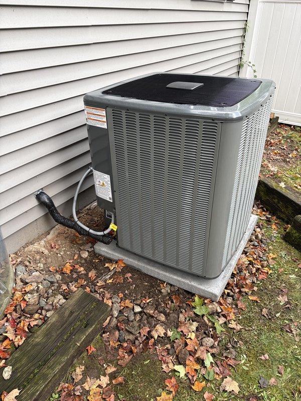 Completed full residential air conditioning system replacement and installation. New outdoor condenser unit installed on existing concrete pad against exterior wall with proper clearances maintained, refrigerant lines connected via black flexible conduit, and all electrical connections secured to manufacturer specifications. Unit tested and confirmed operational with all safety labels properly affixed.