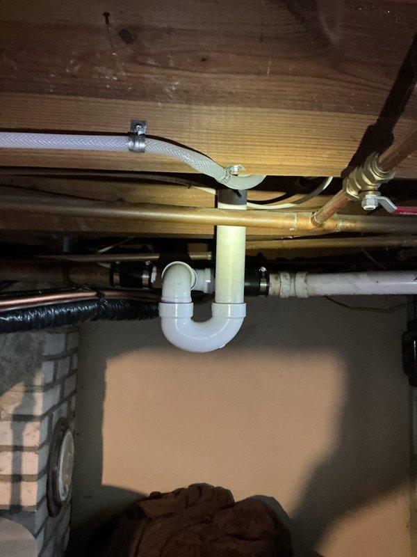 Responded to plumbing leak issue in basement caused by HVAC condensate drain improperly tied into drainage system. Upon inspection, identified that condensate line from Carrier unit was creating overflow conditions in basement whenever plumbing fixtures were flushed. Examined existing P-trap configuration and drainage piping beneath fixtures, noting improper connection between condensate discharge and sanitary waste lines. The condensate drain was causing hydraulic overload in the system, resulting in water backing up and spilling into basement area. Documented plumbing infrastructure including white PVC drain assemblies, P-trap installation, and existing pipe routing along floor joists. Photographed affected areas showing drain trap configuration, copper supply lines, and corrugated condensate hose attachment points to identify source of overflow problem and recommend proper condensate drain routing separate from sanitary waste system.