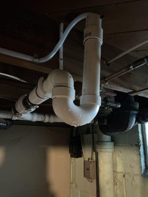 Responded to plumbing leak issue in basement caused by HVAC condensate drain improperly tied into drainage system. Upon inspection, identified that condensate line from Carrier unit was creating overflow conditions in basement whenever plumbing fixtures were flushed. Examined existing P-trap configuration and drainage piping beneath fixtures, noting improper connection between condensate discharge and sanitary waste lines. The condensate drain was causing hydraulic overload in the system, resulting in water backing up and spilling into basement area. Documented plumbing infrastructure including white PVC drain assemblies, P-trap installation, and existing pipe routing along floor joists. Photographed affected areas showing drain trap configuration, copper supply lines, and corrugated condensate hose attachment points to identify source of overflow problem and recommend proper condensate drain routing separate from sanitary waste system.