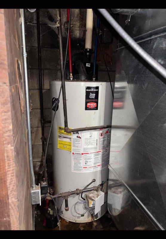 Responded to service call for non-operational electric tank water heater approximately 10 years old. Upon arrival, conducted visual inspection of unit installed in basement utility area between concrete block walls. Assessed water heater condition, noting manufacturer labels intact and multiple pipe connections at top including PVC, copper, and supply lines. Observed floor staining around base of unit indicating previous moisture issues. Examined surrounding area revealing significant water damage to walls and flooring, corroded floor drain with visible deterioration, and evidence of long-term moisture exposure in confined space. Documented equipment condition and installation environment with photographs for service records. Unit accessible but installed in tight quarters with limited clearance. Evaluated electrical connections and plumbing configuration to determine operational status and recommend appropriate corrective action.