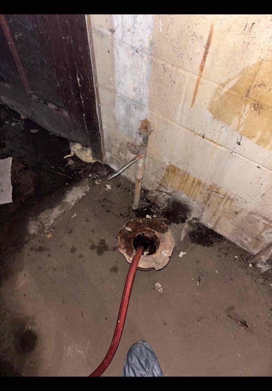 Responded to service call for non-operational electric tank water heater approximately 10 years old. Upon arrival, conducted visual inspection of unit installed in basement utility area between concrete block walls. Assessed water heater condition, noting manufacturer labels intact and multiple pipe connections at top including PVC, copper, and supply lines. Observed floor staining around base of unit indicating previous moisture issues. Examined surrounding area revealing significant water damage to walls and flooring, corroded floor drain with visible deterioration, and evidence of long-term moisture exposure in confined space. Documented equipment condition and installation environment with photographs for service records. Unit accessible but installed in tight quarters with limited clearance. Evaluated electrical connections and plumbing configuration to determine operational status and recommend appropriate corrective action.