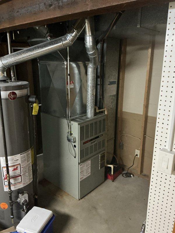 Conducted furnace estimate for club member at operational system scheduled for midday appointment. Documented existing HVAC equipment in basement utility area, including current furnace unit with standard ventilation configuration and associated ductwork system. Photographed equipment layout showing furnace positioned adjacent to water heater with metallic ductwork, insulated horizontal and vertical ducts, and proper venting connections in unfinished basement space with concrete flooring and exposed structural elements.