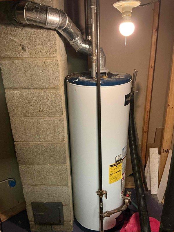 Responded to service call for leaking water heater in basement utility area. Upon inspection, identified active leak on existing white cylindrical unit with blue top and expansion tank assembly. Evaluated plumbing connections, including metallic supply lines, PVC discharge piping, and shut-off valves. Documented current installation showing signs of deterioration with visible moisture, staining, and drip patterns on tank surface. Provided estimate for complete water heater replacement due to compromised tank integrity.