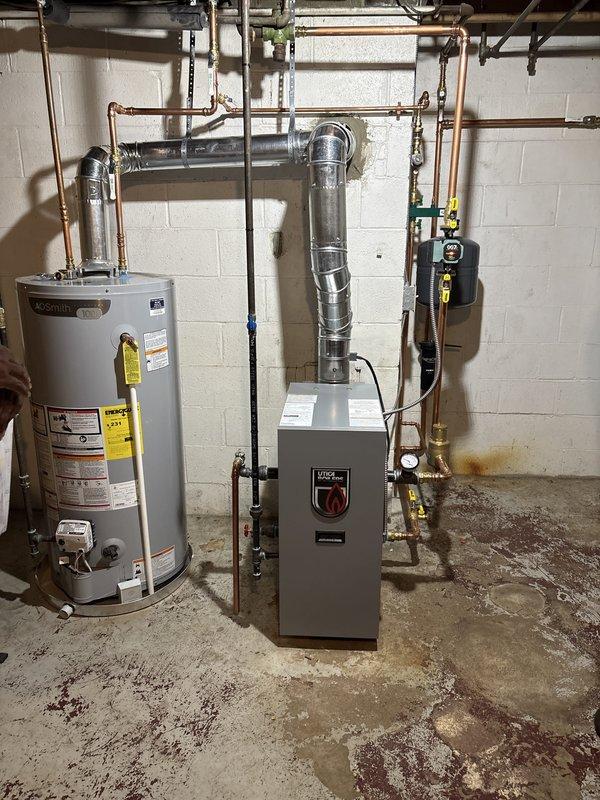 Completed full boiler installation in basement mechanical room, including new gray boiler unit with aluminum venting system routed through ceiling. Installed AO Smith 100-gallon water heater with copper piping connections, pressure relief valves, and proper venting configuration. All equipment mounted against concrete block wall with electrical controls, safety switches, and associated plumbing connections completed per manufacturer specifications.