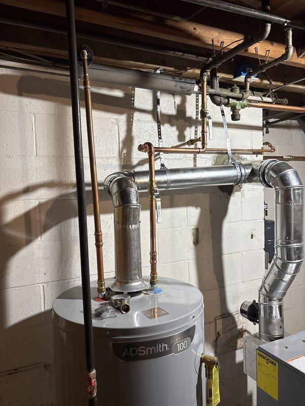Completed full boiler installation in basement mechanical room, including new gray boiler unit with aluminum venting system routed through ceiling. Installed AO Smith 100-gallon water heater with copper piping connections, pressure relief valves, and proper venting configuration. All equipment mounted against concrete block wall with electrical controls, safety switches, and associated plumbing connections completed per manufacturer specifications.