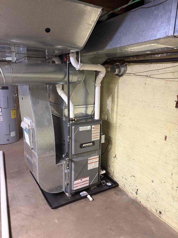 Completed full furnace installation in basement mechanical room. New gray furnace unit was installed on black protective pad with proper PVC venting routed to exterior and metallic ductwork connected to existing distribution system. All equipment properly secured, labeled, and tested for safe operation alongside existing water heater.
