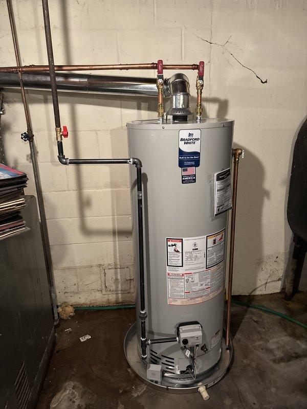 Completed installation of Bradford White residential gas-fired water heater in basement utility room. Installed unit with proper copper piping connections, red shut-off valves on supply lines, and black exhaust vent pipe. Configured gas control assembly and burner access components according to manufacturer specifications. Verified all safety labels, technical specifications, and regulatory compliance information are properly displayed on unit. Installation meets clearance requirements and industry standards for residential gas water heating equipment.