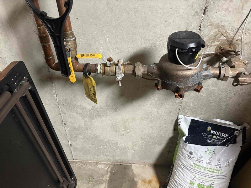 Responded to plumbing leak located at main water meter assembly before the house shut-off valve. Coordinated water shut-off at street level to facilitate repair. Inspected copper piping connections on both sides of the meter and identified source of minor leak at fitting. Repaired compromised connection, restored water service, and verified system integrity with no active leaks present upon completion. Documented existing plumbing configuration including ball valve with yellow handle, water meter with protective cap, and copper supply lines showing age-related patina.