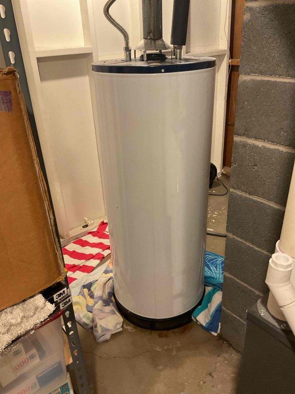 Responded to emergency service call for leaking water heater with water supply shut off. Upon arrival, inspected 17-year-old residential water heater located in basement utility area showing active water leakage at base. Unit exhibited age-related deterioration and compromised tank integrity consistent with end of serviceable life expectancy. Documented existing installation configuration including gas connections, venting system, and plumbing connections. Photographed water heater positioning between wooden framing and concrete block walls, noting accessible clearances and utility room layout. Provided estimate for complete water heater replacement including removal of existing failed unit and installation of new equipment to restore hot water service to residence.
