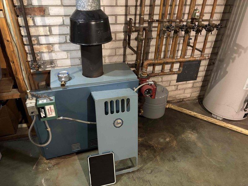 Responded to a boiler complaint of an abnormal (non-blue) burner flame and failure to maintain set temperature on a cast-iron, gas-fired hydronic boiler of approximately 50 years in age. Inspected the unit, venting, and associated components, including the draft hood and flue, circulator pump, expansion tank, and control and wiring connections, and observed floor staining and discoloration around the base of the boiler and aged copper piping joints with soldering residue. Based on the unit's age, combustion and performance issues, and visible signs of deterioration, discussed replacement options with the homeowner.