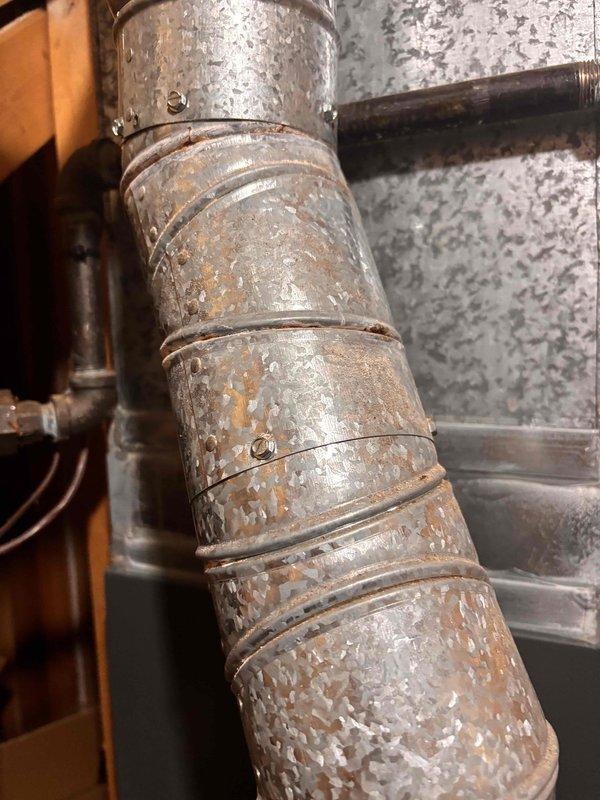 Performed annual heating tune-up on 22-year-old Bryant furnace and inspected the galvanized flue vent pipe for the surface crack reported by the gas company. Observed scattered surface corrosion and oxidation along the vent pipe sections, particularly near the band joints and seams, with no obvious separation or major damage visible; connections remain secured with metal bands and fasteners. Recommend continued monitoring of the vent pipe condition and consideration of vent replacement as the corrosion progresses.