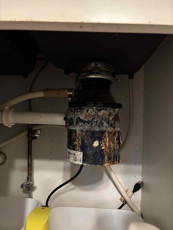 Performed annual furnace tune-up on the EnergiAir system, inspecting and servicing components for safe, efficient operation; no humidifier is installed. Photos taken during the visit documented a heavily corroded garbage disposal under the kitchen sink, with rust staining, mineral buildup, and a small hole in the housing, indicating a likely leak and need for replacement.