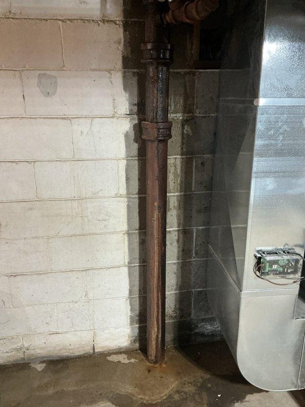 Inspected the reported sewer line leak and found active moisture at a corroded cast iron drain stack in the basement, with wetting and rust residue at the pipe couplings and pooling at the base of the stack. This is a recurring condition following prior partial drain repipe and P-trap work. The affected section of drain line remains a source of leakage, and replacement of that section is recommended to permanently resolve the issue.