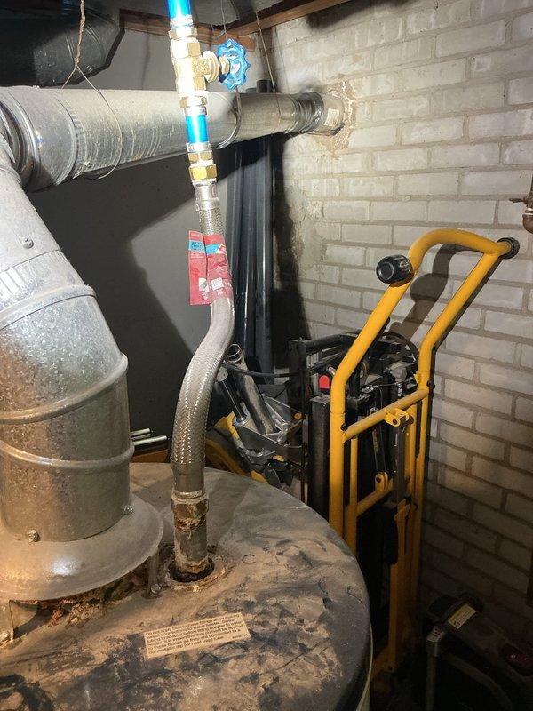 Inspected the water heater and identified corrosion and rust staining at the braided stainless steel flexible water connector fitting on the tank's top. Evaluated the cold-water supply connection, brass shutoff valve, and PEX transition, and noted dust accumulation on the tank, deteriorated mortar where the galvanized vent connector meets the masonry wall, and rust-colored sealant staining at the adjacent plenum seams. Documented all conditions with photographs for the service record.
