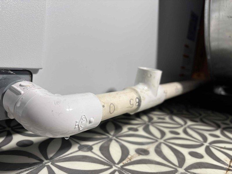 Responded to a recurring water leak at the AprilAire 600A humidifier and inspected the PVC drain assembly at the base of the equipment cabinet. Found active dripping at the PVC elbow fitting and moisture residue at the adjacent tee fitting, with no cracks, rust, or frost observed on the cabinet or ductwork. Documented the leaking drain joints with photos and verified the unit's warranty status for follow-up on the repeat failure.