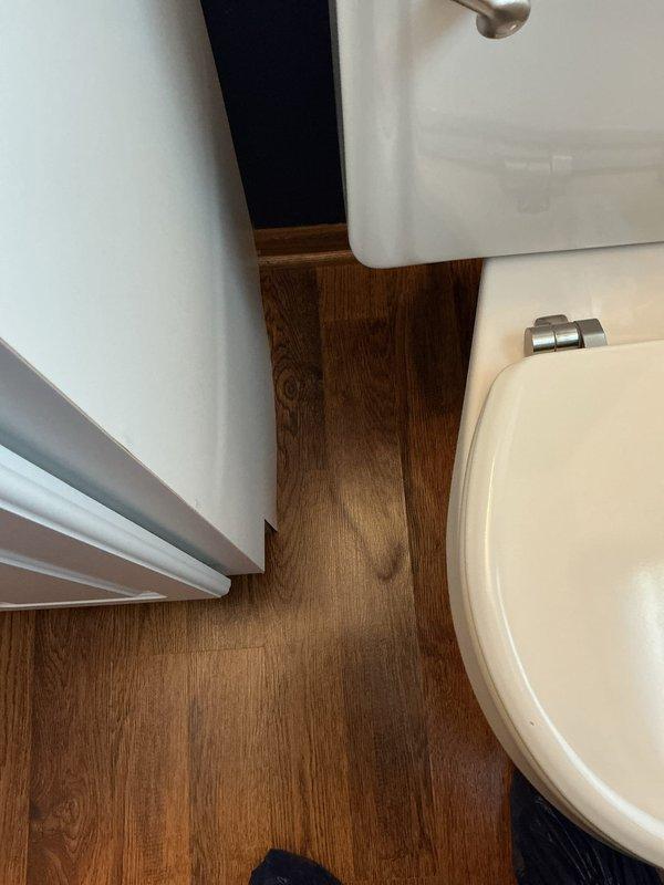 Inspected the reported leak at the base of the toilet and found the bowl was not securely bolted down or sealed to the floor, with plastic shims wedged beneath the front edge and discoloration on the flooring adjacent to the base. Address of the wax seal failure and unsecured fixture was indicated, requiring the toilet to be reset with a new wax seal and secured to the flange to eliminate the leak.