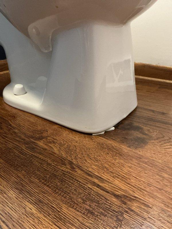 Inspected the reported leak at the base of the toilet and found the bowl was not securely bolted down or sealed to the floor, with plastic shims wedged beneath the front edge and discoloration on the flooring adjacent to the base. Address of the wax seal failure and unsecured fixture was indicated, requiring the toilet to be reset with a new wax seal and secured to the flange to eliminate the leak.