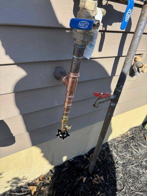 Performed on-site evaluation of an exterior hose bib and backflow preventer assembly to assess relocation of the outside faucet line to facilitate irrigation system drain-down. Inspected the existing brass backflow preventer with dual ball isolation valves, noting a weathered, tarnished supply line alongside newly installed copper piping with a tee fitting and hose-bib outlet. Documented existing conditions with photographs and evaluated routing options to establish scope of work for the proposed relocation.