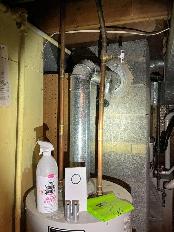 Responded to a reported leak at an 11-year-old tank-style water heater and inspected the unit, associated copper supply piping, shutoff valve, and galvanized flue vent routed through the masonry wall. Documented the condition of the vent penetration, which has a deteriorated cement patch, as well as the items stored on top of the tank, and evaluated the unit for the source of the leak.