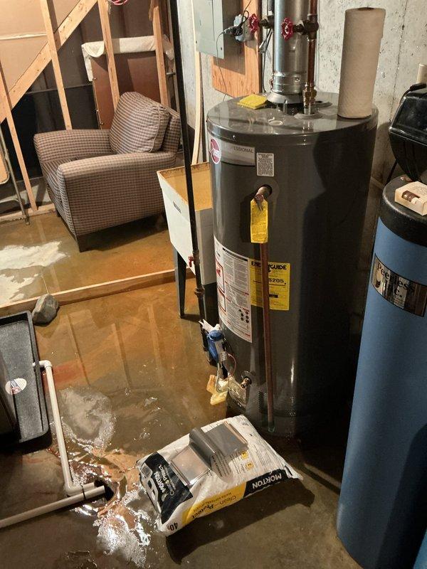 Performed an on-site estimate for replacement of an aging (15+ year) Rheem tank-style water heater located in a utility area with standing water present on the floor. Inspected the water heater, flue, shutoff valves, and adjacent water softener and ductwork, and documented existing conditions and equipment data labels with photographs to support replacement quoting.