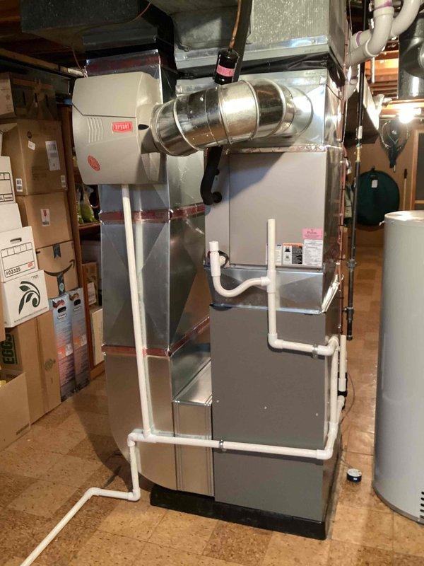 Completed installation of new residential gas furnace in basement mechanical room. Installed vertical air handler unit with proper ductwork connections, integrated humidifier system with water supply lines, and white PVC condensate drain piping with appropriate support brackets. All equipment mounted securely on basement floor with professional routing of drainage lines, electrical connections, and sealed metallic duct joints at supply plenum.