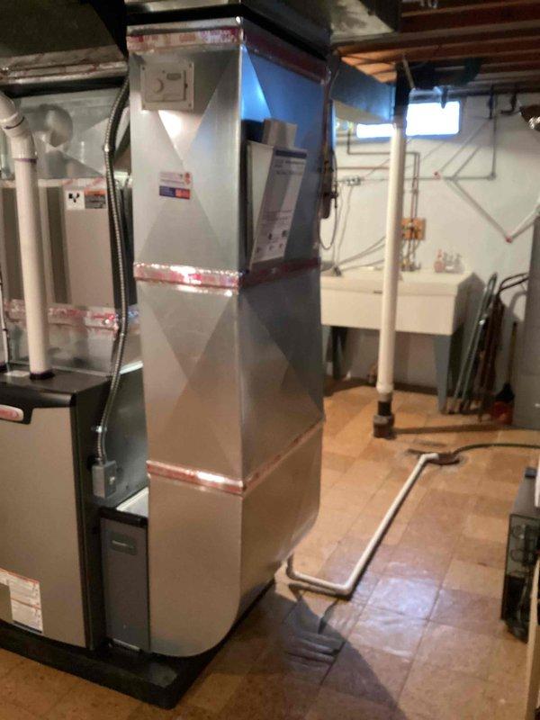 Completed installation of new residential gas furnace in basement mechanical room. Installed vertical air handler unit with proper ductwork connections, integrated humidifier system with water supply lines, and white PVC condensate drain piping with appropriate support brackets. All equipment mounted securely on basement floor with professional routing of drainage lines, electrical connections, and sealed metallic duct joints at supply plenum.