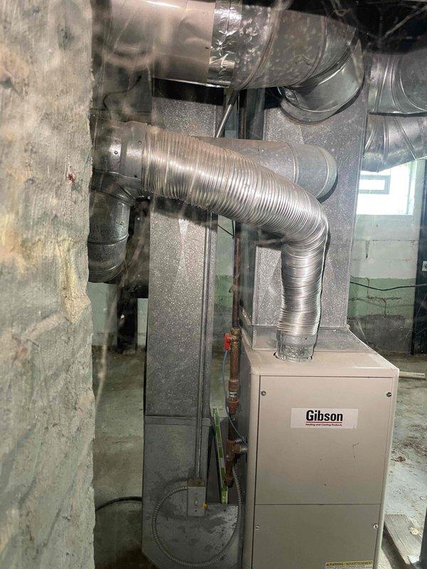 Conducted comprehensive site evaluation for full HVAC system replacement in residential property currently lacking air conditioning. Assessed existing 15-year-old Gibson furnace installation in basement utility area, documenting current heating infrastructure including extensive flexible aluminum ductwork network with multiple branches and elbows throughout ceiling space. Evaluated existing duct connections, metal clamps, and tape sealing at junction points. Photographed furnace unit with two-tone gray and beige cabinet configuration, cylindrical supply plenum, and associated gas line and electrical connections. Inspected concrete block foundation walls, basement layout constraints, and available space for new equipment installation. Provided detailed estimate for complete HVAC system installation including new central air conditioning unit to meet customer's timeline of replacement within the next couple weeks.