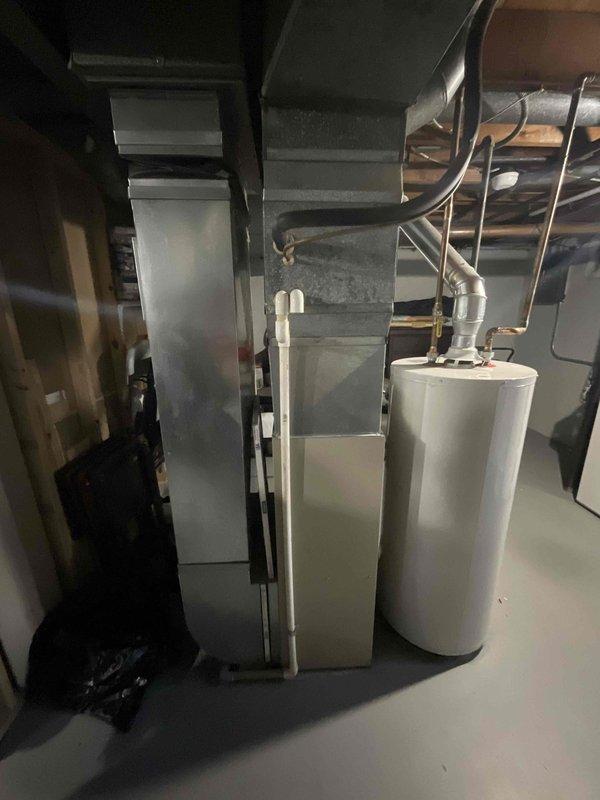Conducted comprehensive site evaluation for complete HVAC system replacement on residential property with 16-year-old air conditioning system. Assessed existing exterior condenser unit positioned on concrete pad against brick exterior wall, noting beige/tan unit with protective metal grille and mounted electrical disconnect. Evaluated interior equipment including vertical air handler/furnace unit and water heater in basement utility area, documenting ductwork configuration, clearances, and mechanical connections. Photographed both exterior and interior installations to document current system layout, equipment condition, and spatial requirements for proposed full system replacement scheduled within the next couple of weeks.