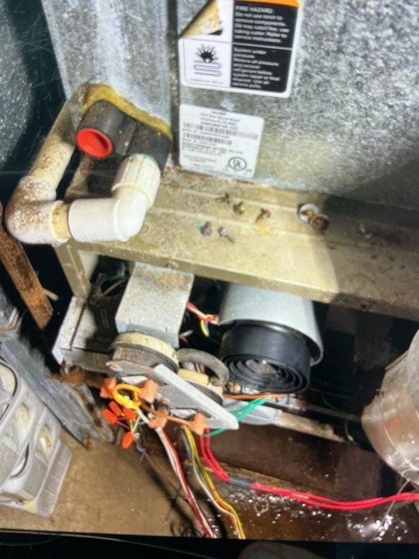 Responded to no-heat call on 15-year-old forced air furnace system that had experienced flooding. Upon arrival, conducted thorough inspection of unit and surrounding components. Found extensive corrosion and oxidation on metal piping throughout the system, indicating prolonged water exposure and moisture damage. Documented internal components including water heater tank, mounting brackets, PVC piping with connections, and electrical wiring assembly. Assessed condition of all visible components including motor, pump unit, and electrical connections with wire nuts. Photographed corroded vertical pipes displaying heavy rust and oxidation patterns, as well as internal mechanical systems showing dust accumulation and age-related wear. System confirmed non-operational at time of inspection due to flood damage.