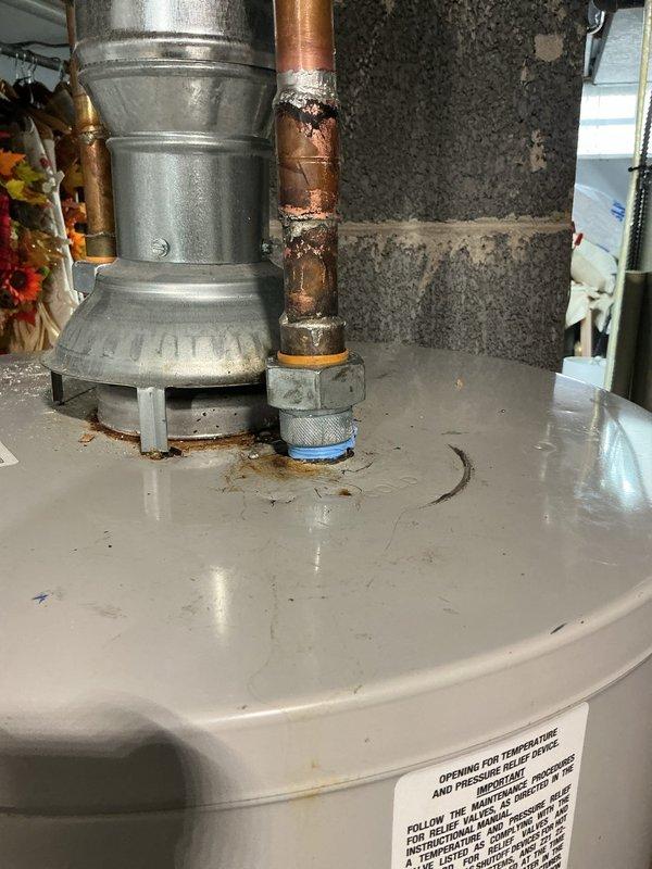 Responded to service call for leaking water heater located in basement. Upon inspection, identified active leak at top connection point where copper supply pipe meets tank through brass fitting on 2012 model tanked water heater. Visual evidence confirmed water droplets present at pipe junction, with corrosion and oxidation visible on copper piping and fittings. Performed diagnostic evaluation of unit and documented leak source with photographic evidence showing moisture accumulation at connection point and deteriorated condition of plumbing connections.