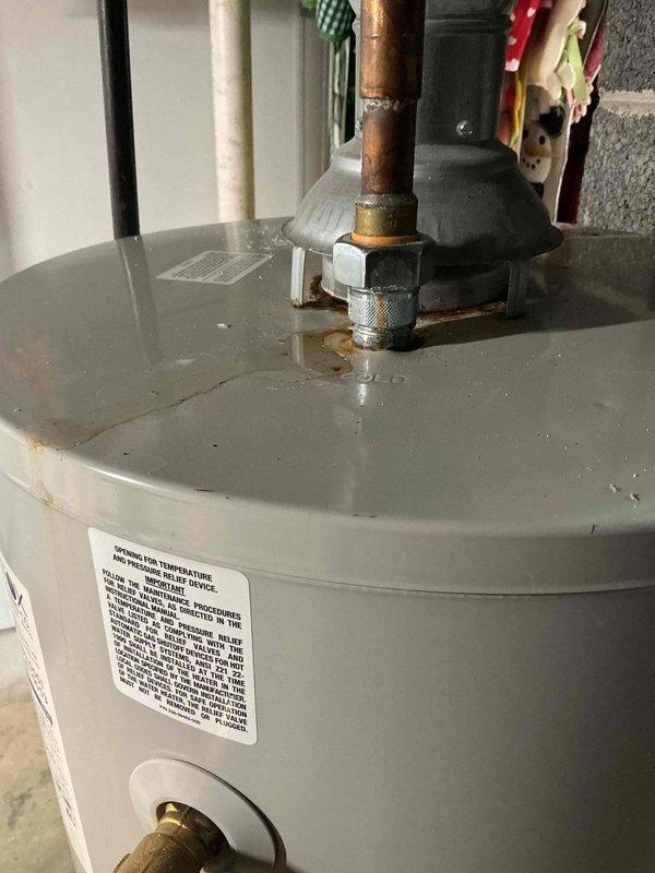 Responded to service call for leaking water heater located in basement. Upon inspection, identified active leak at top connection point where copper supply pipe meets tank through brass fitting on 2012 model tanked water heater. Visual evidence confirmed water droplets present at pipe junction, with corrosion and oxidation visible on copper piping and fittings. Performed diagnostic evaluation of unit and documented leak source with photographic evidence showing moisture accumulation at connection point and deteriorated condition of plumbing connections.