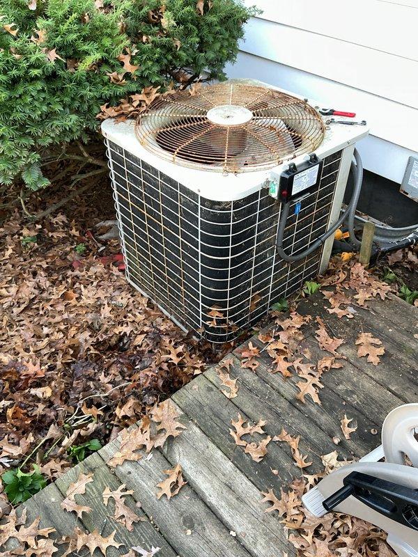 Completed annual preventative maintenance on residential air conditioning condenser unit. Performed comprehensive system inspection including electrical testing with multimeter showing proper voltage readings at 12.3V, verified fan motor operation and inspected protective grille showing normal weathering. Cleared accumulated debris and leaves from around unit and decking area to ensure proper airflow and drainage. All components operating within normal parameters upon completion.