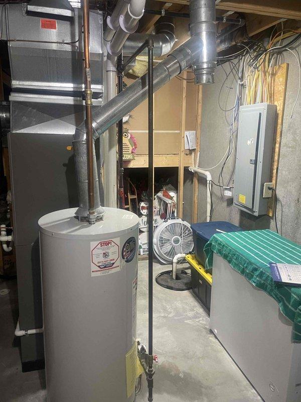 Responded to service call for leaking water heater in residential basement. Upon inspection, confirmed active leak from original water heater installed in 2007, indicating unit has reached end of serviceable life after approximately 18 years of operation. Documented existing equipment specifications and measurements, noting standard cylindrical tank configuration with overhead black metal flue pipe connection and brass drain valve at base. Assessed surrounding mechanical room infrastructure including proximity to HVAC system, electrical panel, and existing plumbing connections. Photographed current installation setup showing water heater positioning relative to concrete walls, overhead ductwork, and utility access points. Measured tank dimensions to determine replacement specifications and clearance requirements for new unit installation. Evaluated venting configuration and gas line connections to ensure compliance with current code requirements for replacement equipment. Prepared comp