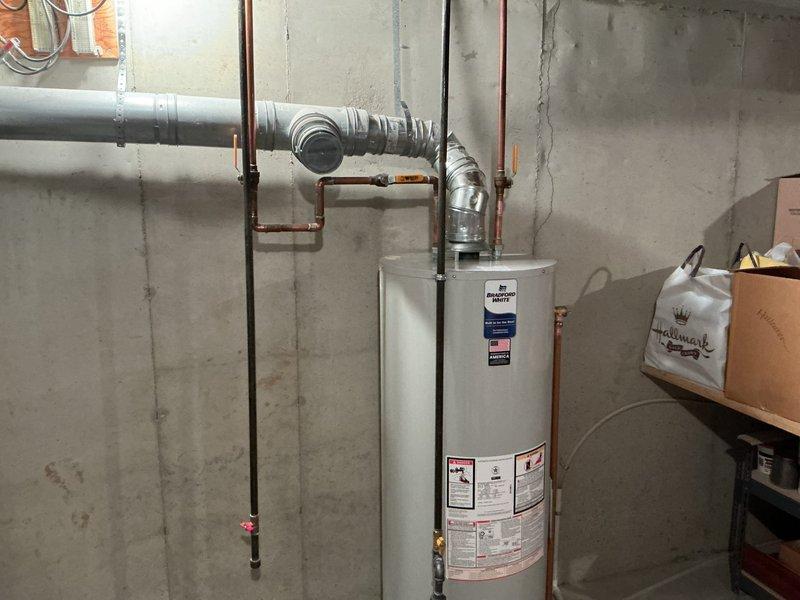 Completed water heater replacement in basement utility area. Removed existing unit and installed new white cylindrical water heater with proper venting connections, including metallic exhaust vent pipe with flexible ribbed ducting connected to rigid metal ductwork. Verified all copper pipe connections and ensured system integration with existing plumbing infrastructure.