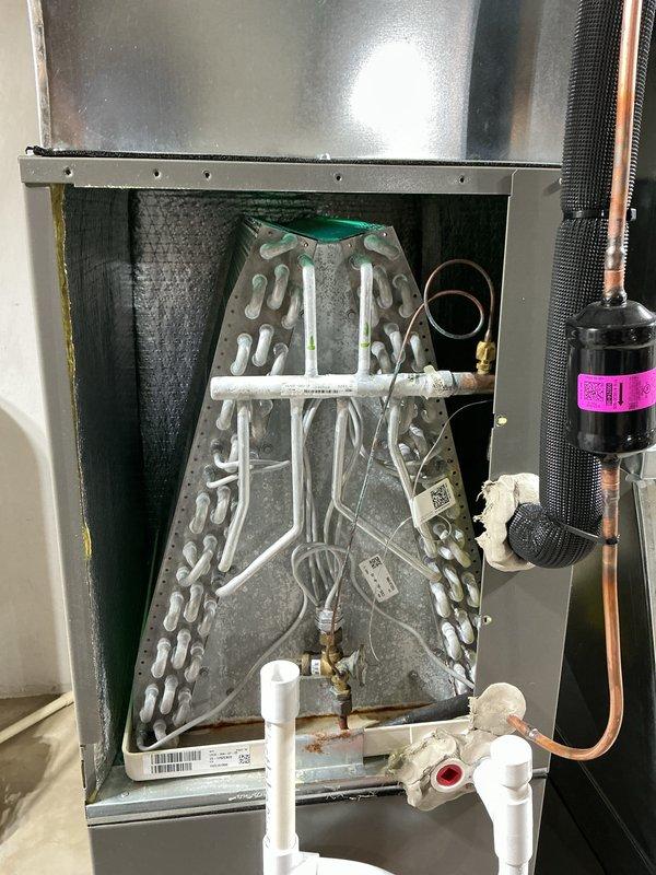 Completed annual cooling maintenance on HVAC air handler unit. Accessed interior cabinet and inspected A-frame evaporator coil assembly, noting light dust accumulation on serpentine tubing surfaces. Verified refrigerant line connections on suction and liquid lines with proper copper line integrity. Inspected filter drier component with manufacturer labeling intact and properly mounted. Checked condensate drain system with white PVC piping in place and functioning. Replaced air filter with clean pleated media filter showing proper accordion-fold construction and structural support framework. All electrical connections and wiring inspected and found secure. Unit operational and ready for cooling season.