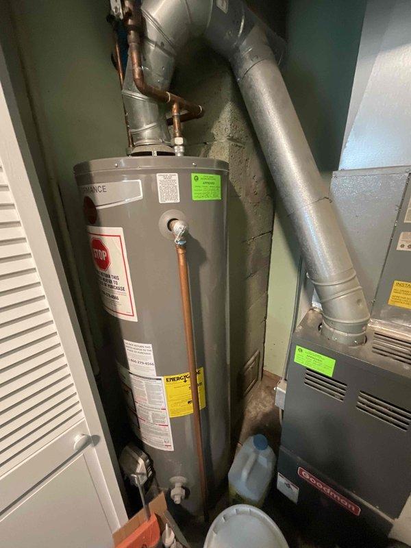 Conducted on-site evaluation of existing 15-year-old furnace and air conditioning system for replacement estimate. Inspected gray furnace unit located in utility closet with vertical paneling, noting manufacturer information and current operating condition. Assessed ductwork configuration including silver metal ducts extending from furnace top. Evaluated water heater with copper piping connections and associated plumbing infrastructure in mechanical room. Documented equipment placement in compact utility space with adjacent storage areas. Photographed current HVAC system installation, including furnace labels, ventilation grilles, and duct routing along wall surfaces. Reviewed system age and operational status to provide comprehensive replacement recommendations for both heating and cooling equipment.