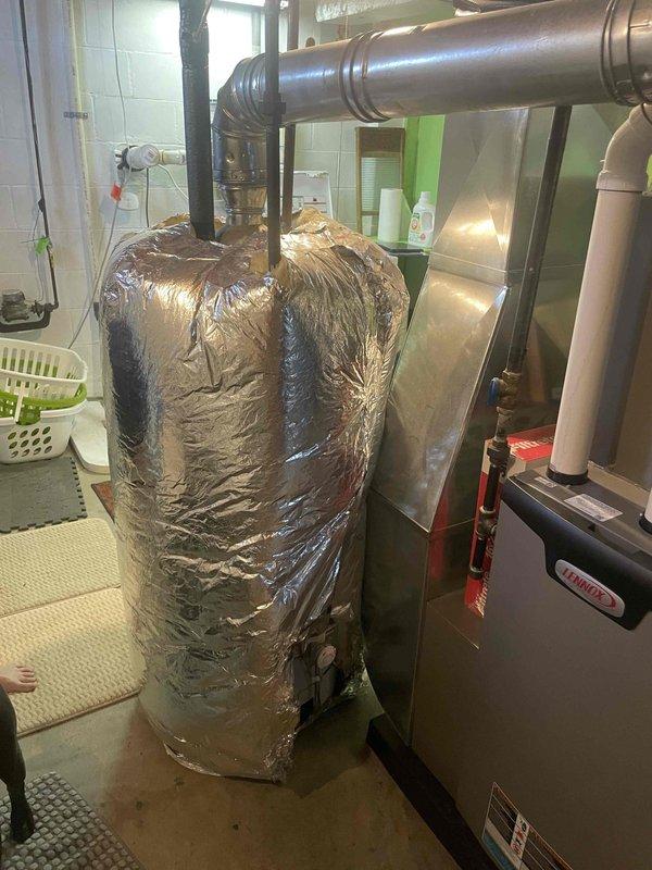 Conducted on-site evaluation of existing water heater system for replacement estimate. Assessed current unit configuration including insulated tank, pipe connections, venting arrangement, and clearance requirements in mechanical room. Documented equipment specifications, installation conditions, and spatial constraints to develop comprehensive replacement proposal. Photographed existing water heater with reflective insulation wrap, black supply/discharge pipes, and proximity to HVAC equipment to ensure accurate sizing and compatibility recommendations for new unit installation.