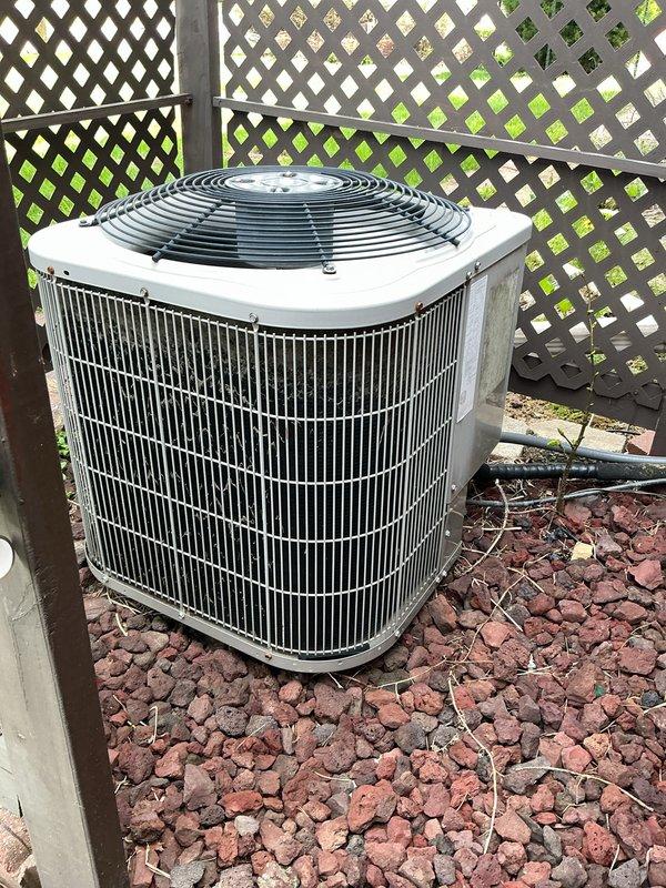 Arrived on-site for scheduled annual HVAC maintenance on residential air conditioning condensing unit. Performed comprehensive visual inspection of outdoor condenser unit, which is positioned on concrete pad with lava rock landscaping in corner location behind lattice fencing. Unit exterior and fan guard showed good overall condition with minimal weathering. Verified condenser coil fins are intact and accessible for airflow. Documented equipment condition with photographs for service records. Unit operational and maintaining proper function at time of inspection.