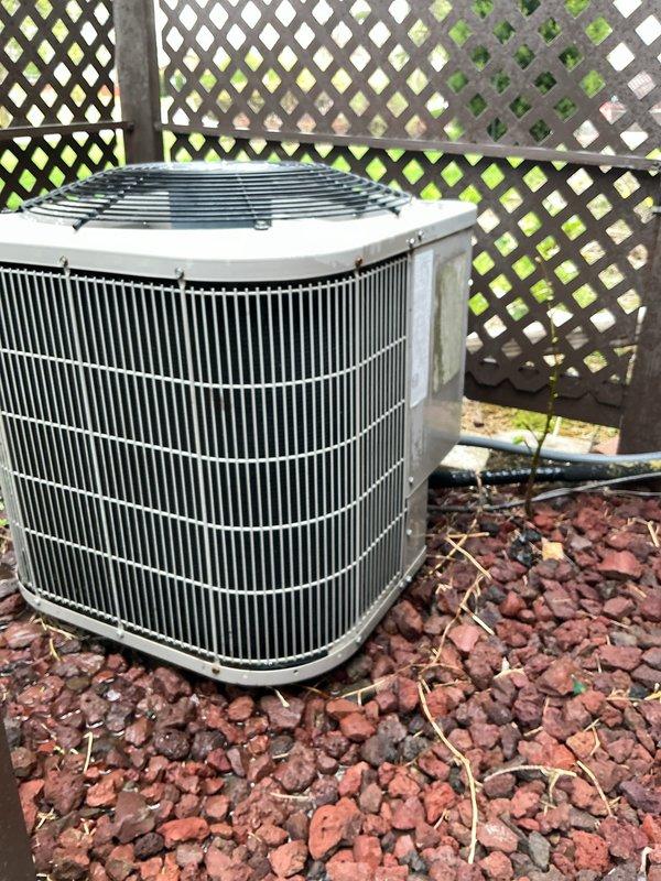 Arrived on-site for scheduled annual HVAC maintenance on residential air conditioning condensing unit. Performed comprehensive visual inspection of outdoor condenser unit, which is positioned on concrete pad with lava rock landscaping in corner location behind lattice fencing. Unit exterior and fan guard showed good overall condition with minimal weathering. Verified condenser coil fins are intact and accessible for airflow. Documented equipment condition with photographs for service records. Unit operational and maintaining proper function at time of inspection.