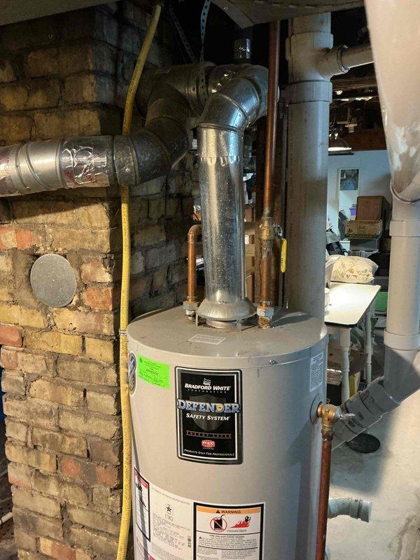 Responded to no hot water complaint affecting entire household. Upon arrival, inspected Bradford White Defender Safety System tanked water heater located in basement utility area. Conducted diagnostic evaluation of unit including gas supply line, pilot light assembly, thermocouple, and burner operation. Verified proper venting through metallic flue pipe system and checked all copper water line connections for integrity. Assessed yellow flexible gas line routing along brick wall for code compliance and safety. Tested temperature and pressure relief valve functionality and examined unit for signs of sediment buildup or internal tank failure. Documented existing installation conditions including PVC drainage system, exposed utilities, and overall water heater setup in century-old home environment. Restored hot water service to all fixtures throughout residence.