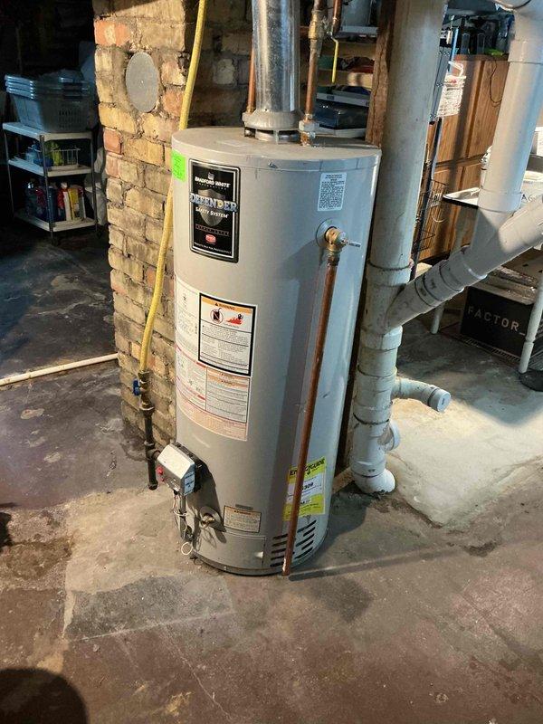 Responded to no hot water complaint affecting entire household. Upon arrival, inspected Bradford White Defender Safety System tanked water heater located in basement utility area. Conducted diagnostic evaluation of unit including gas supply line, pilot light assembly, thermocouple, and burner operation. Verified proper venting through metallic flue pipe system and checked all copper water line connections for integrity. Assessed yellow flexible gas line routing along brick wall for code compliance and safety. Tested temperature and pressure relief valve functionality and examined unit for signs of sediment buildup or internal tank failure. Documented existing installation conditions including PVC drainage system, exposed utilities, and overall water heater setup in century-old home environment. Restored hot water service to all fixtures throughout residence.