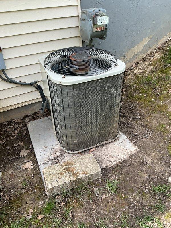 Completed annual cooling maintenance on residential outdoor condenser unit. Inspected cylindrical condenser housing, verified fan operation through protective grille, and assessed overall unit condition. Noted weathering and dirt accumulation on metal grille surfaces and housing requiring cleaning attention. Confirmed electrical connections at disconnect box and conduit routing along building exterior remain secure. Unit positioned on concrete pad with proper clearances maintained. Documented high-efficiency filter spots requiring monitoring during next service interval.