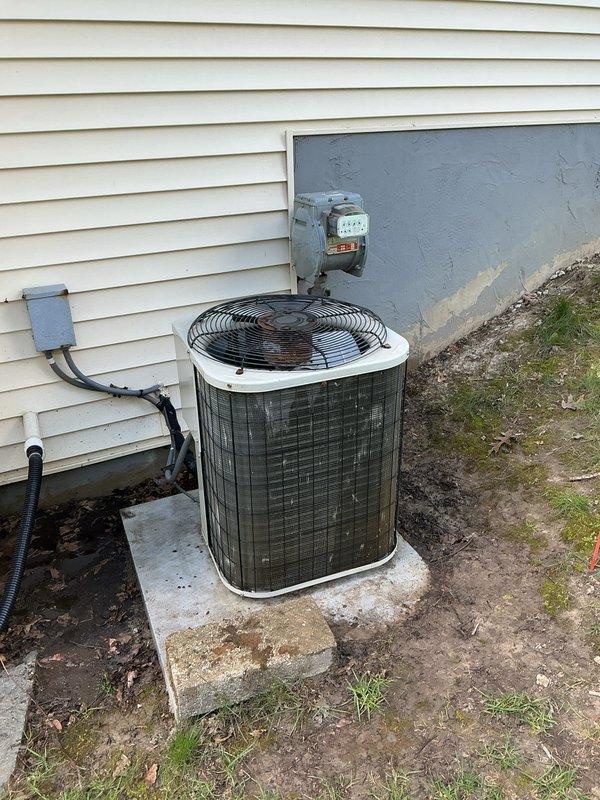 Completed annual cooling maintenance on residential outdoor condenser unit. Inspected cylindrical condenser housing, verified fan operation through protective grille, and assessed overall unit condition. Noted weathering and dirt accumulation on metal grille surfaces and housing requiring cleaning attention. Confirmed electrical connections at disconnect box and conduit routing along building exterior remain secure. Unit positioned on concrete pad with proper clearances maintained. Documented high-efficiency filter spots requiring monitoring during next service interval.