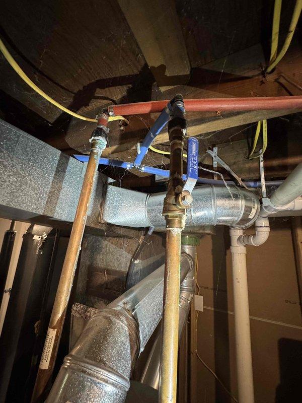 Responded to service call for reported leak and low water temperature issues at tankless water heater installation. Upon inspection, identified water accumulation in utility area beneath plumbing system. Examined tankless water heater connections, supply lines, and associated piping including copper supply lines, PEX distribution pipes, and isolation valves. Assessed all visible connections, fittings, and valve assemblies for signs of active leakage or seepage. Documented existing plumbing configuration including main supply connections with blue-handled isolation valves, branch piping, and ductwork proximity to water lines. Photographed installation for service records showing overhead utility space layout and mechanical equipment arrangement.