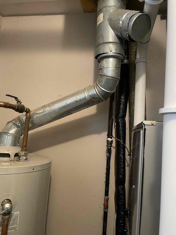 Conducted on-site assessment for water heater replacement in utility area. Inspected existing white cylindrical water heater unit, documenting current installation configuration including copper flex connector, galvanized ductwork connections, and associated plumbing lines. Measured tank dimensions and evaluated surrounding infrastructure including black insulated refrigerant lines, PVC drainage, and wall-mounted components. Photographed existing setup showing T-junction ductwork, mounting brackets, and connection points to provide accurate estimate for hot water heater replacement. Unit showed signs of age with dust accumulation on top surface. Assessment completed to prepare comprehensive replacement quote.